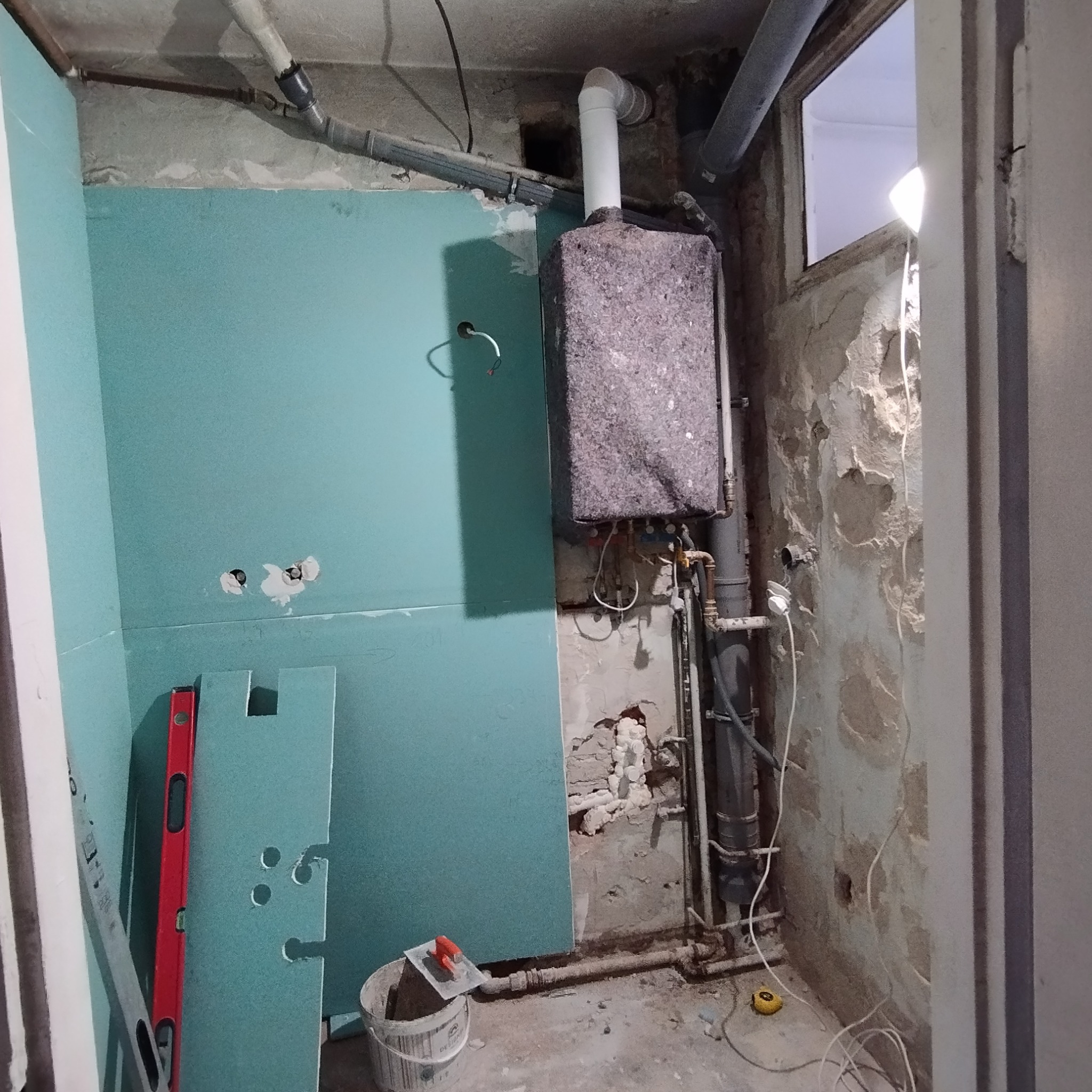 Remontowana łazienka: płyty gipsowe na ścianach, instalacja grzewcza, rury, poziomica i wiadro z narzędziami. Etap prac wykończeniowych.