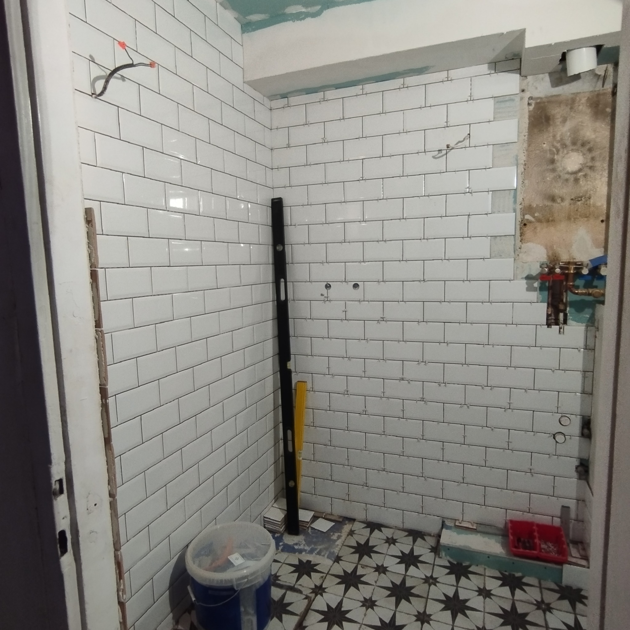 Układanie białych, prostokątnych płytek w łazience, widoczny poziomica, wiadro i fragment instalacji. Podłoga z wzorem gwiazd.