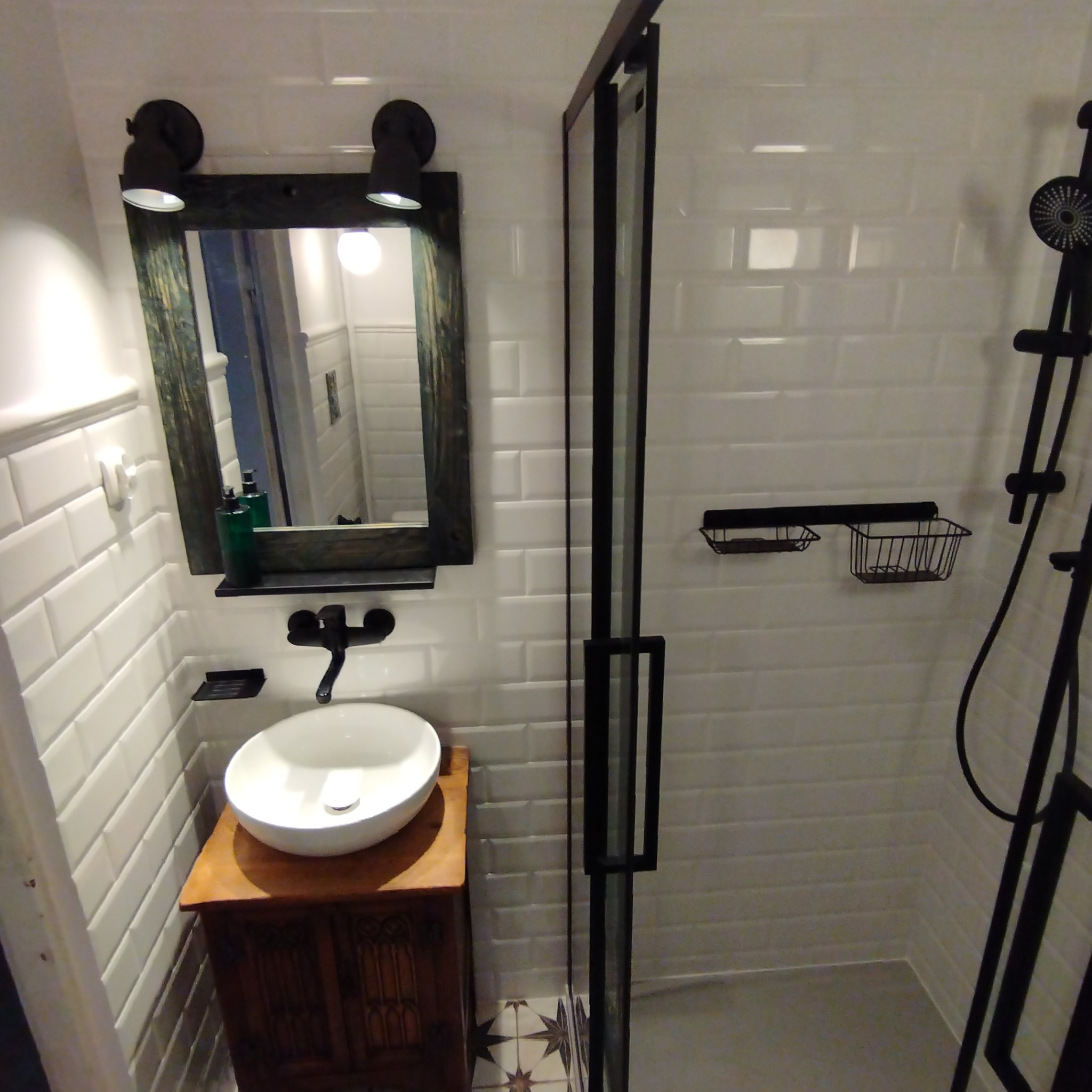 Nowoczesna łazienka z białą, cegiełkową glazurą, kabiną prysznicową z czarnymi ramami i umywalką na drewnianej szafce. Widoczne czarne akcenty i oświetlenie.