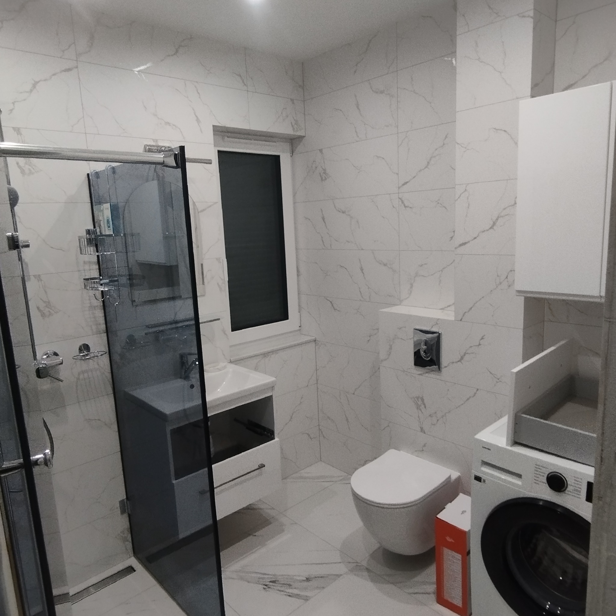 Nowoczesna łazienka z kabiną prysznicową typu walk-in, umywalką z szafką, toaletą podwieszaną i pralką. Białe płytki imitujące marmur na ścianach i podłodze.