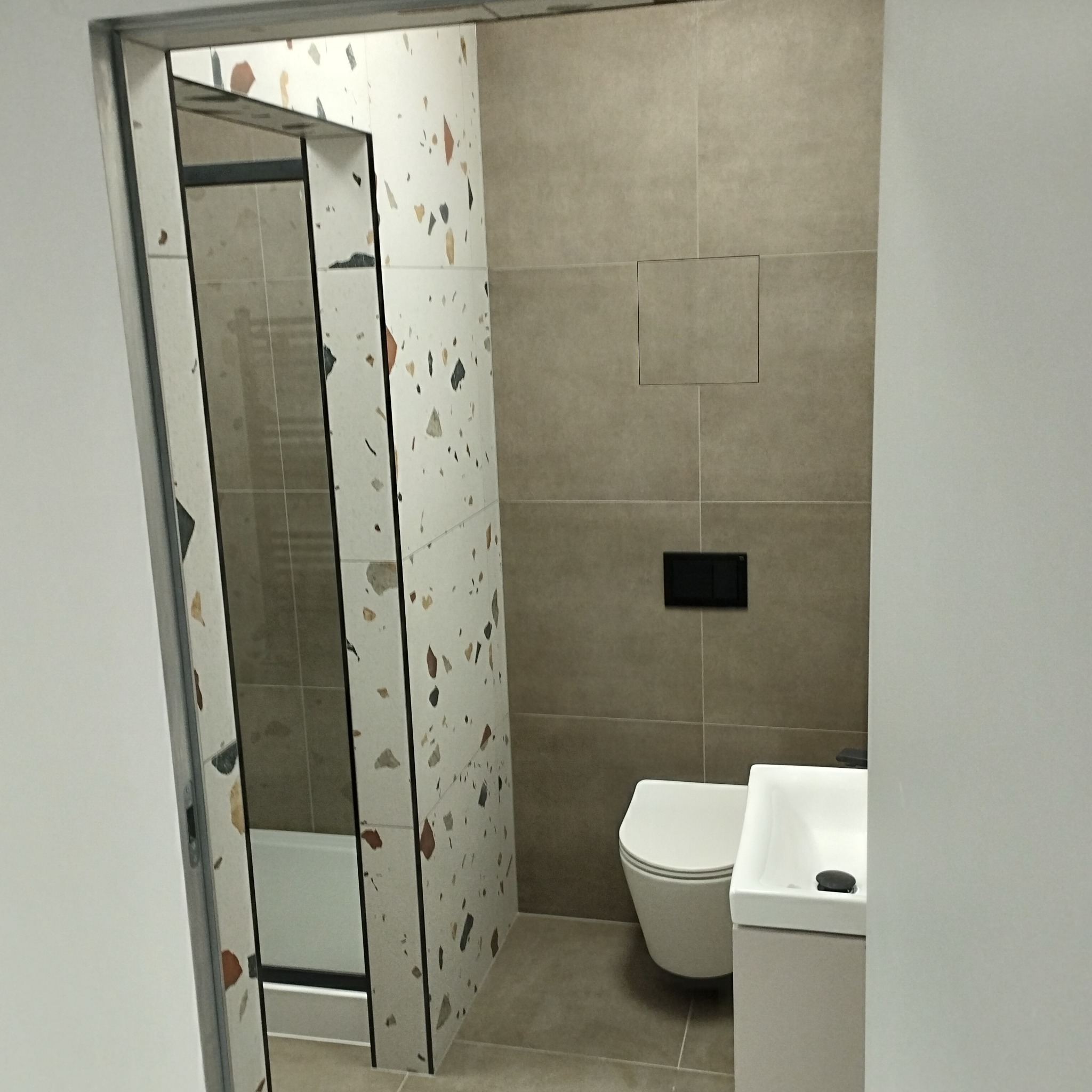 Nowoczesna łazienka z kabiną prysznicową, umywalką i toaletą. Płytki w odcieniach beżu i bieli z kolorowymi akcentami. Minimalistyczny design, czarne detale.