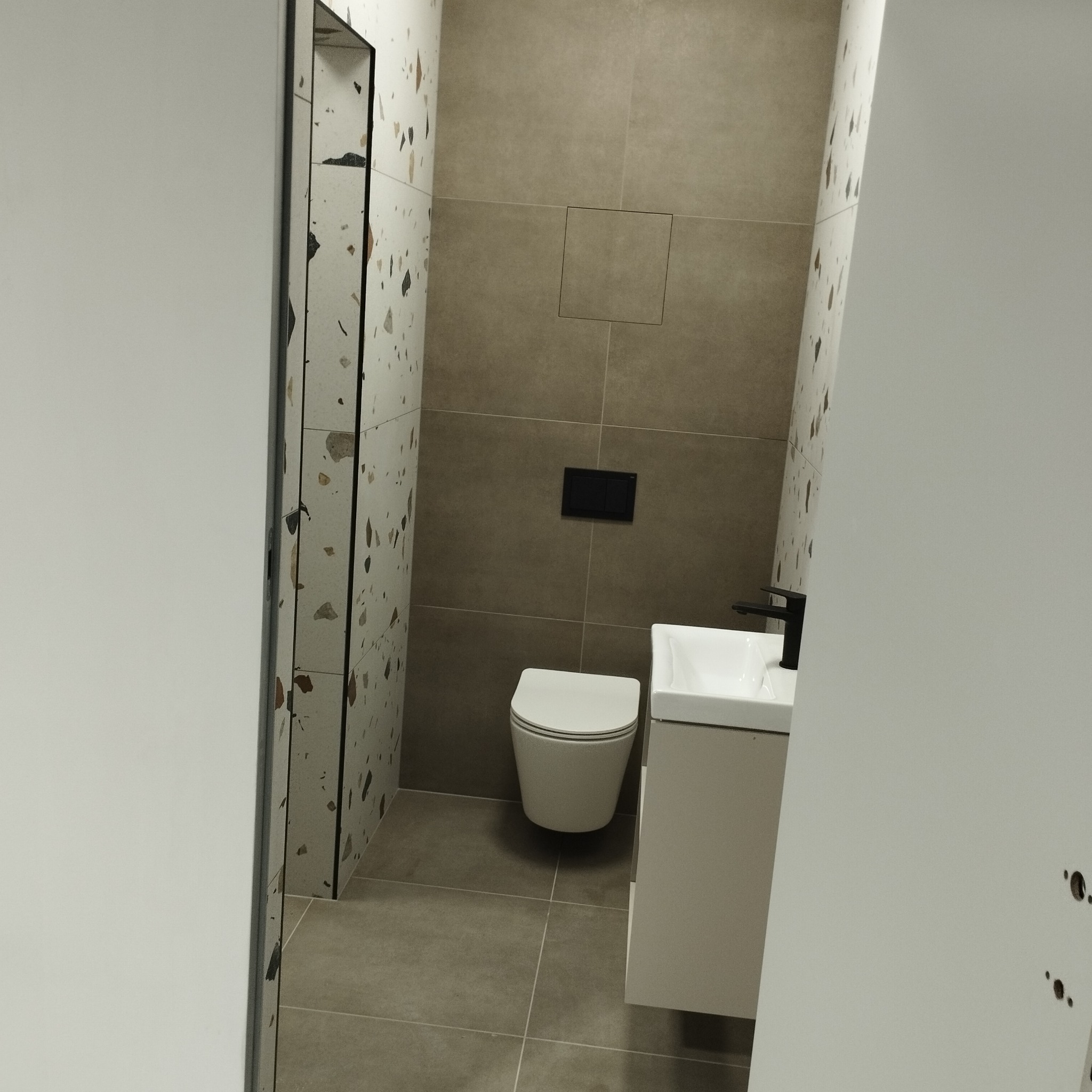 Nowoczesna łazienka w szarościach z umywalką na szafce, toaletą i płytkami imitującymi lastryko. Widok zza uchylonych drzwi.