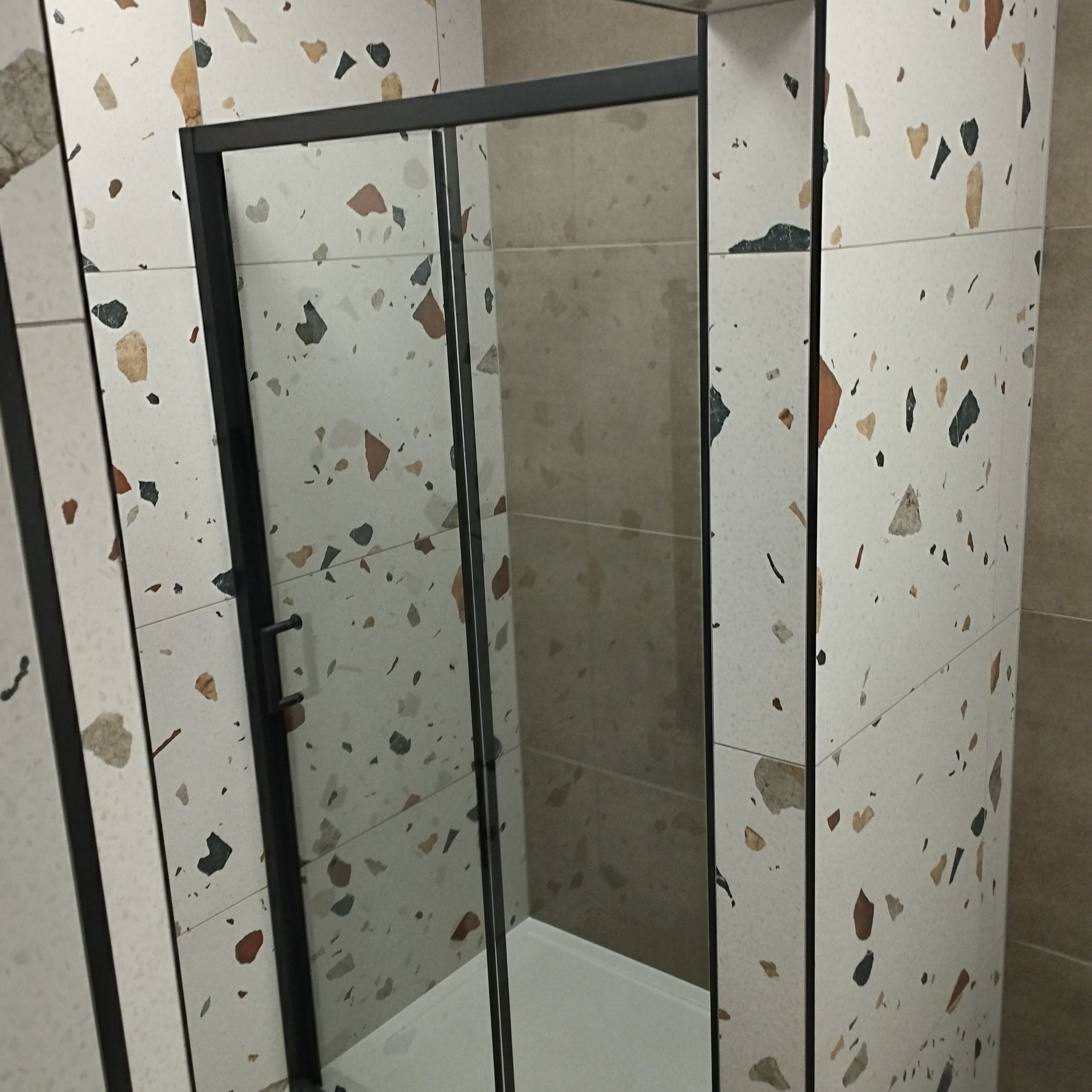 Kabina prysznicowa z czarnymi profilami na tle płytek lastryko w odcieniach bieli i beżu. Widoczny brodzik. Minimalistyczny design, nowoczesne wykończenie łazienki.