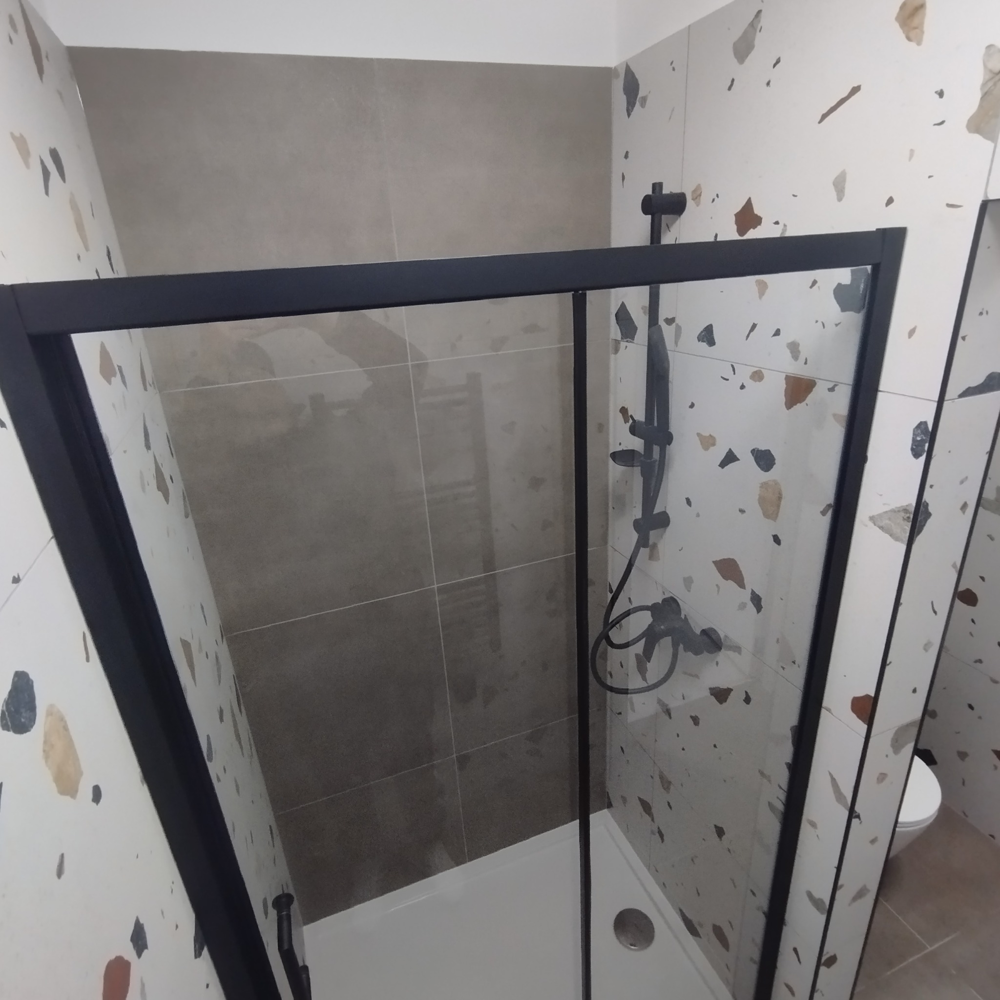 Nowoczesna kabina prysznicowa z czarną ramą, szklanymi drzwiami i deszczownicą. Ściany wyłożone płytkami w dwóch wzorach: gładkie szare i mozaika lastryko.