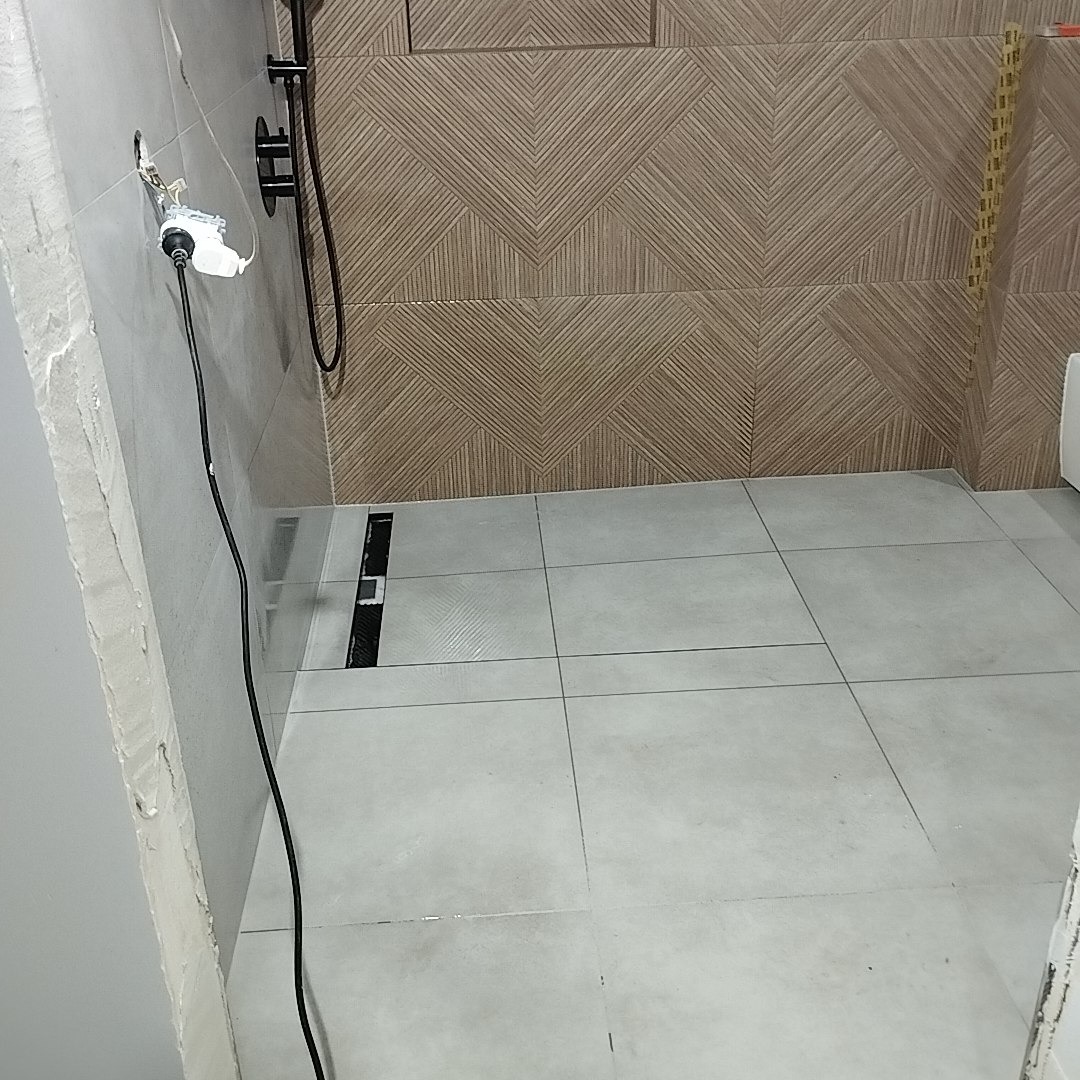 Ukończona łazienka z szarymi płytkami podłogowymi i dekoracyjną ścianą z geometrycznym wzorem. Widoczny odpływ liniowy i fragment instalacji elektrycznej.