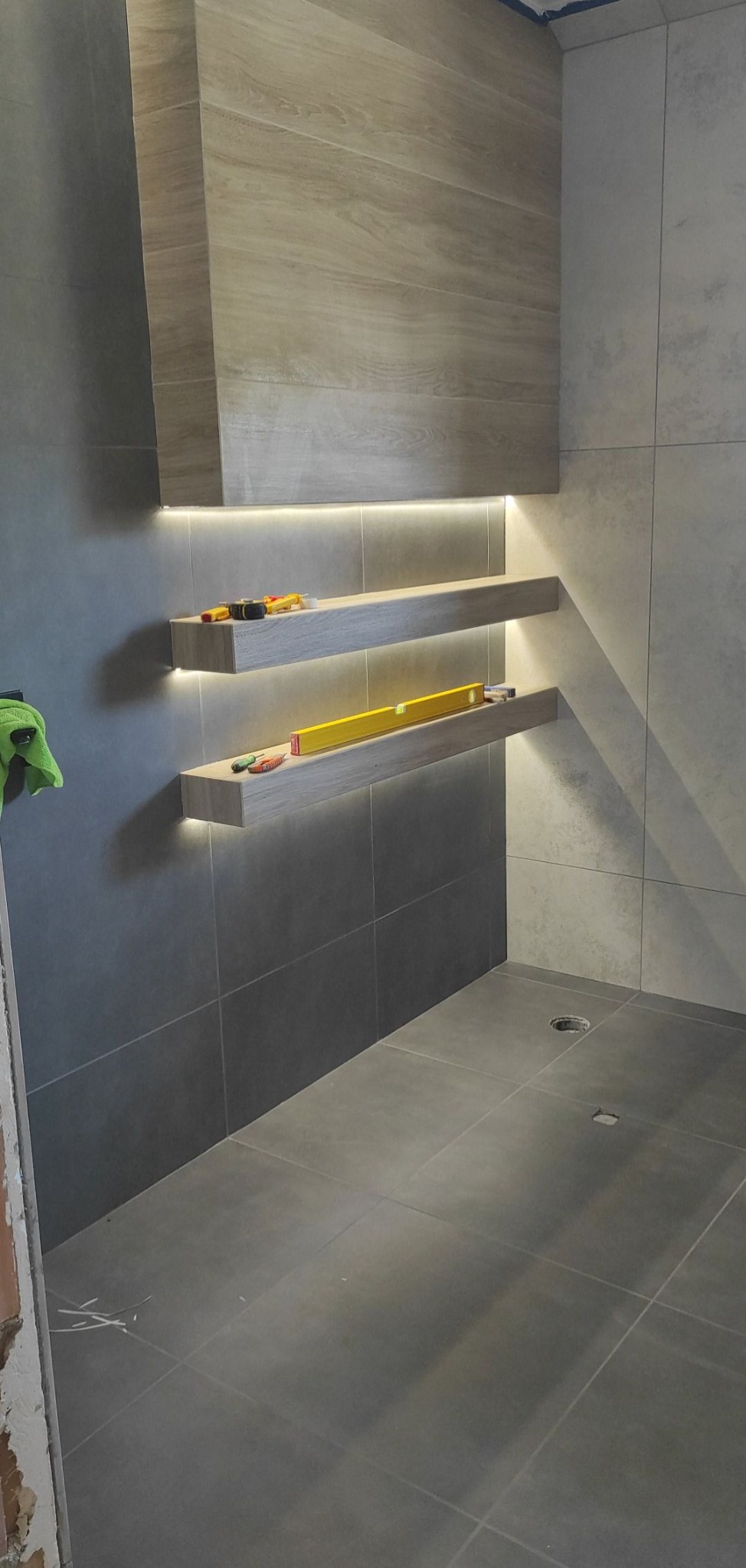 Nowoczesna łazienka z szarymi płytkami i podświetlanymi półkami z drewnopodobnym wykończeniem. Na półkach leżą poziomice i narzędzia.