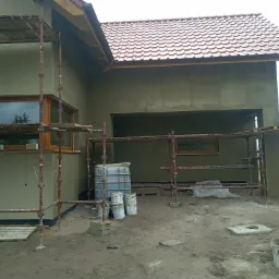 Dom w trakcie prac elewacyjnych, z nałożonym tynkiem, widoczne rusztowania, pojemniki z zaprawą i narzędzia na placu budowy.