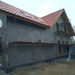 Dom w trakcie ocieplania, widoczne szare płyty izolacyjne na elewacji, rusztowania i częściowo wykończony dach z czerwonej dachówki.