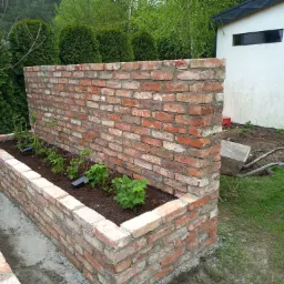 Mur z czerwonej cegły tworzący podwyższoną grządkę z posadzonymi roślinami, na tle trawnika i białego budynku gospodarczego z czarnym oknem.