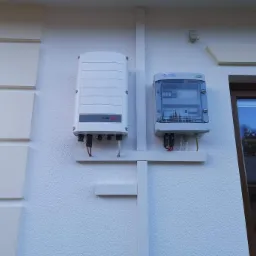 Biały inwerter SolarEdge i skrzynka elektryczna Alfa na ścianie budynku, połączone białymi listwami maskującymi przewody, widok z boku okna.