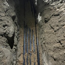 BoWe Energy Jakub Mucha - Wykop z ułożonymi, czarnymi rurami w ziemi, zasypanymi piaskiem. Widoczny fragment buta w dolnym rogu. Ujęcie z góry, ukazujące etap prac ziemnych.