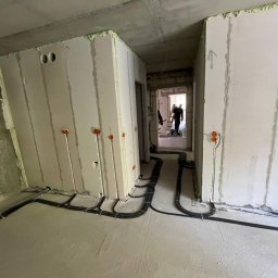 BoWe Energy Jakub Mucha - Wnętrze w trakcie prac elektrycznych: widoczne kable prowadzone po podłodze i wyprowadzone przewody w ścianach z pomarańczowymi puszkami. W tle sylwetka osoby.