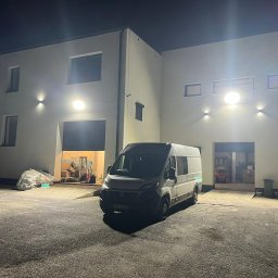 BoWe Energy Jakub Mucha - Biały budynek oświetlony lampami zewnętrznymi, z zaparkowanym vanem na placu. W tle widoczne otwarte wrota garażowe i wózek widłowy.