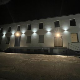 BoWe Energy Jakub Mucha - Oświetlony budynek nocą z równomiernie rozmieszczonymi lampami elewacyjnymi, tworzącymi efektowne stożki światła nad każdym oknem i drzwiami.