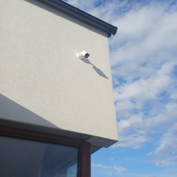 BoWe Energy Jakub Mucha - Biała kamera monitoringu zamontowana na elewacji budynku z widocznym oknem i fragmentem nieba z chmurami. Ujęcie z dołu, podkreślające nowoczesny design.