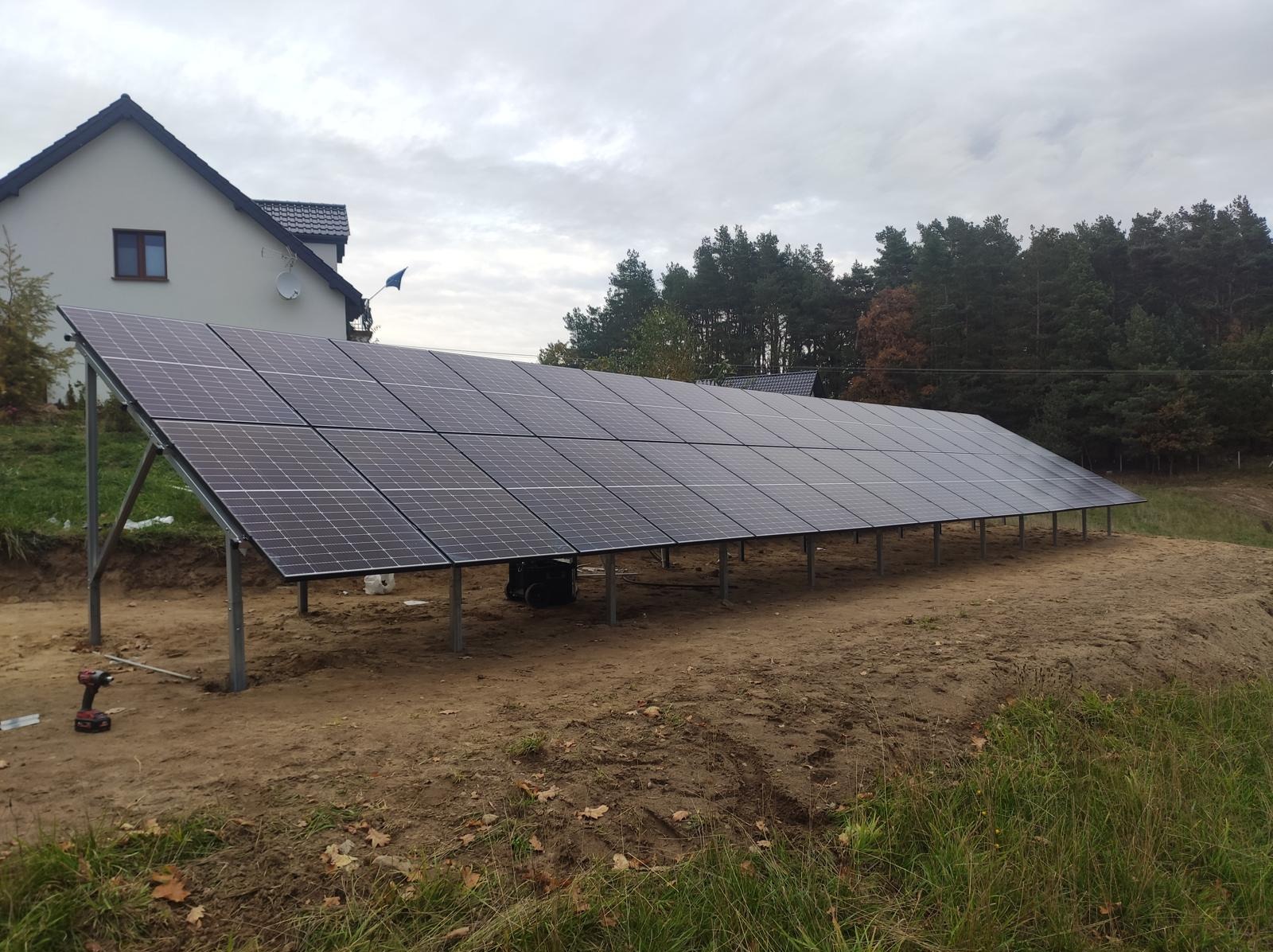 Długa instalacja fotowoltaiczna na metalowej konstrukcji, z widocznym wkrętakiem i torbą narzędziową, na tle domu i lasu w pochmurny dzień.