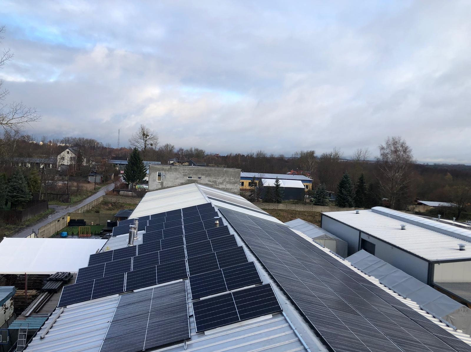 Widok z góry na panele fotowoltaiczne zamontowane na dachu budynku przemysłowego, w tle zabudowa jednorodzinna i pochmurne niebo. Widoczne kominy wentylacyjne.