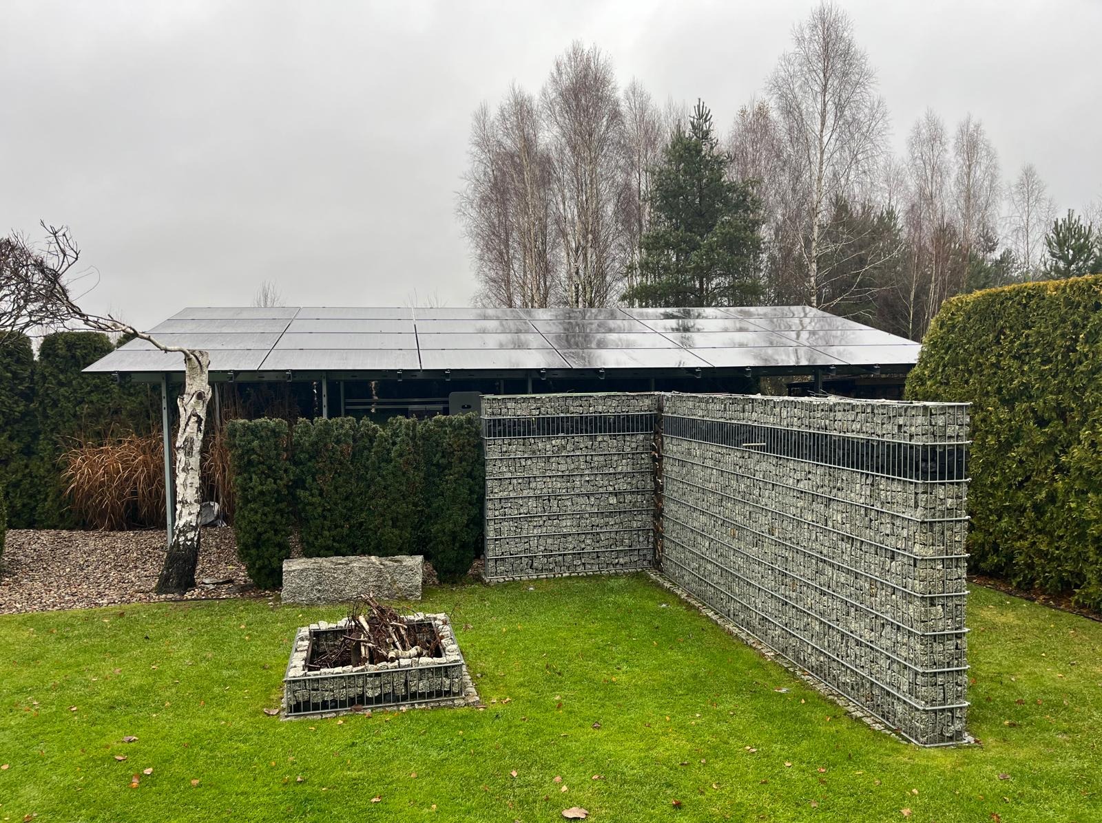 Nowoczesna instalacja fotowoltaiczna na zadaszeniu w ogrodzie, widoczna z perspektywy trawnika. Gabiony tworzą ogrodzenie, a w tle drzewa bez liści i pochmurne niebo.