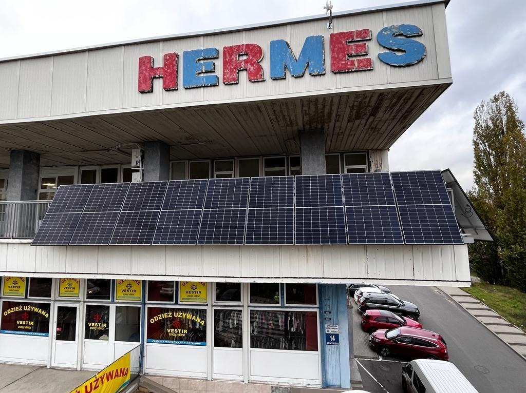 Panele fotowoltaiczne zamontowane na dachu budynku z szyldem 'HERMES' i sklepem odzieżowym na parterze. Widok z zewnątrz, pochmurne niebo w tle.