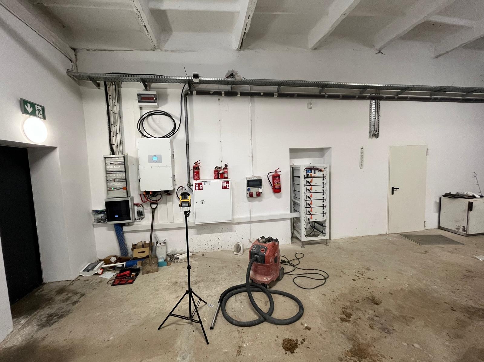 Wnętrze pomieszczenia z zamontowaną instalacją fotowoltaiczną, widoczne panele, inwerter, zabezpieczenia, narzędzia pomiarowe i odkurzacz przemysłowy. Białe ściany i sufit.
