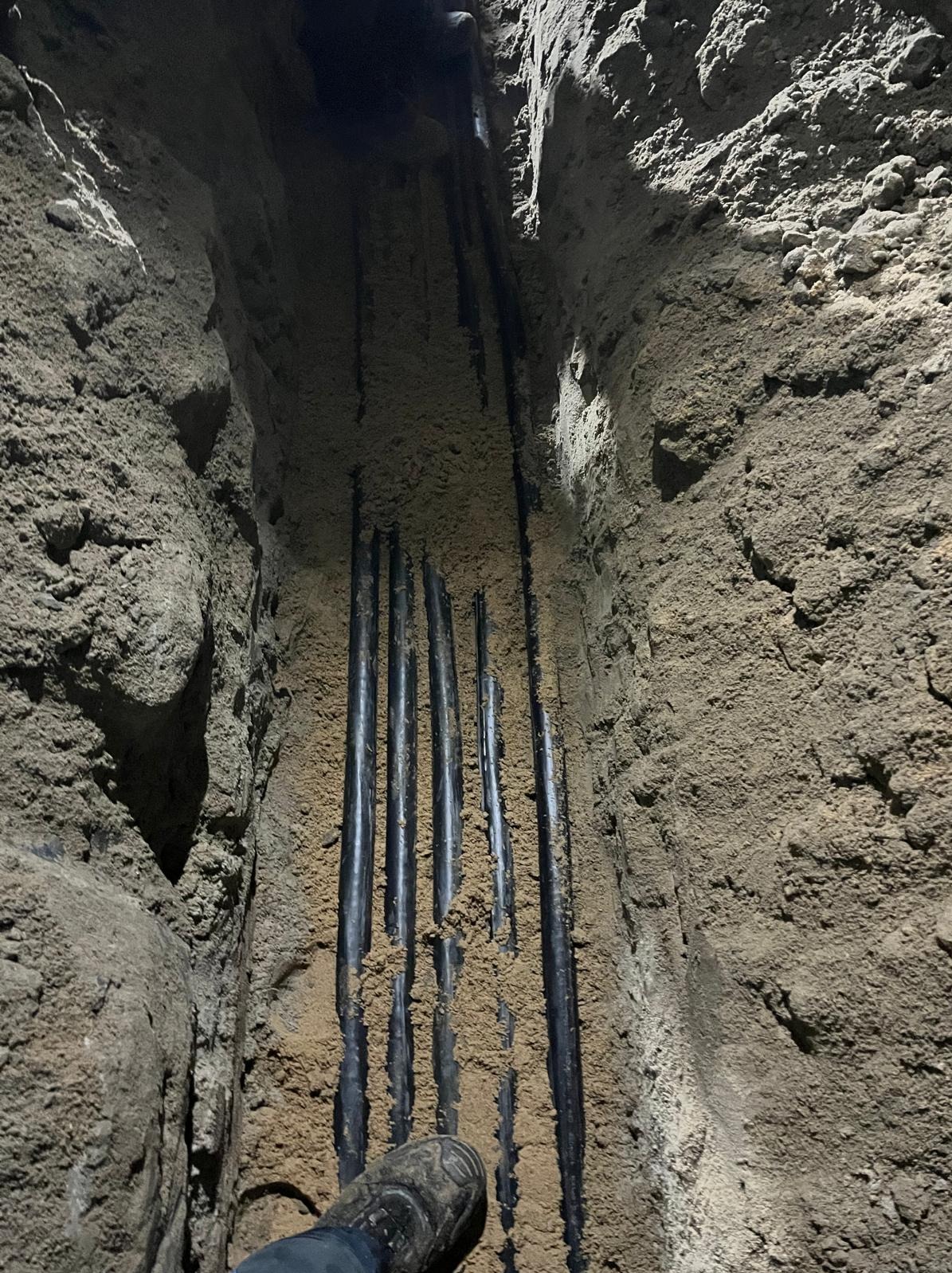 Wykop z ułożonymi, czarnymi rurami w ziemi, zasypanymi piaskiem. Widoczny fragment buta w dolnym rogu. Ujęcie z góry, ukazujące etap prac ziemnych.