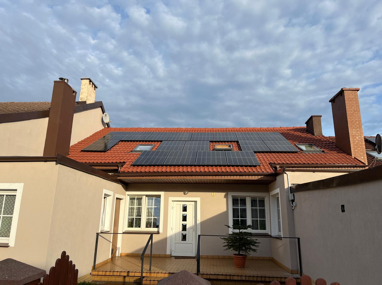 Dom jednorodzinny z panelami fotowoltaicznymi na czerwonym dachu, widok z dołu, błękitne niebo z chmurami w tle. Na pierwszym planie balkon z małą choinką.