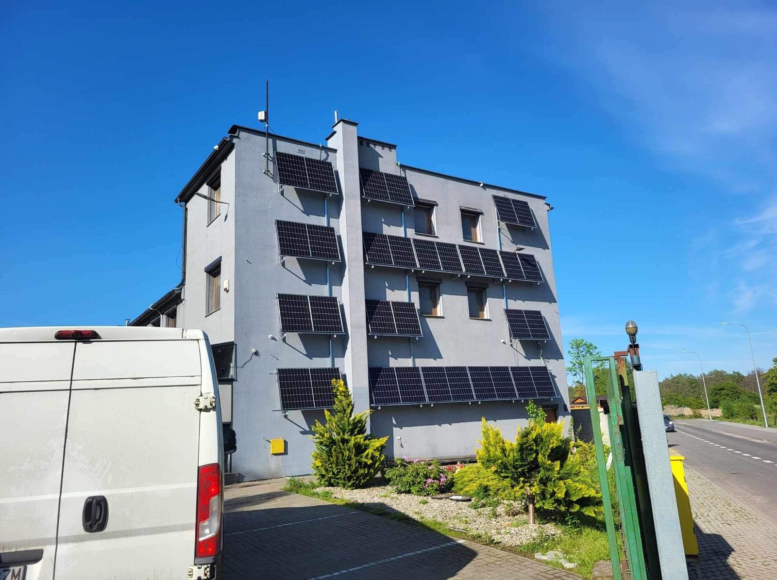 Szary budynek z panelami słonecznymi zamontowanymi na elewacji w Kołczygłowach. Częściowo widoczny biały bus. Błękitne niebo.