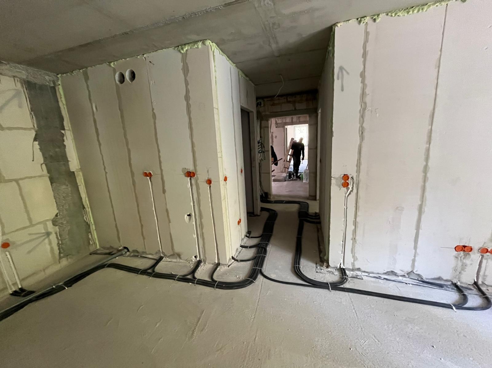 Wnętrze w trakcie prac elektrycznych: widoczne kable prowadzone po podłodze i wyprowadzone przewody w ścianach z pomarańczowymi puszkami. W tle sylwetka osoby.