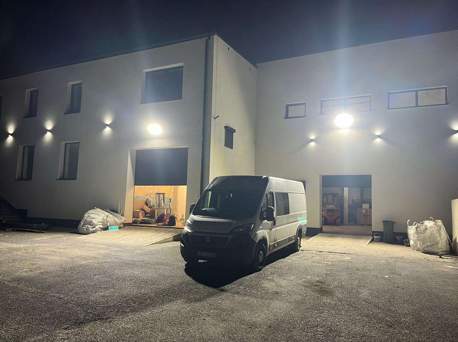 Biały budynek oświetlony lampami zewnętrznymi, z zaparkowanym vanem na placu. W tle widoczne otwarte wrota garażowe i wózek widłowy.