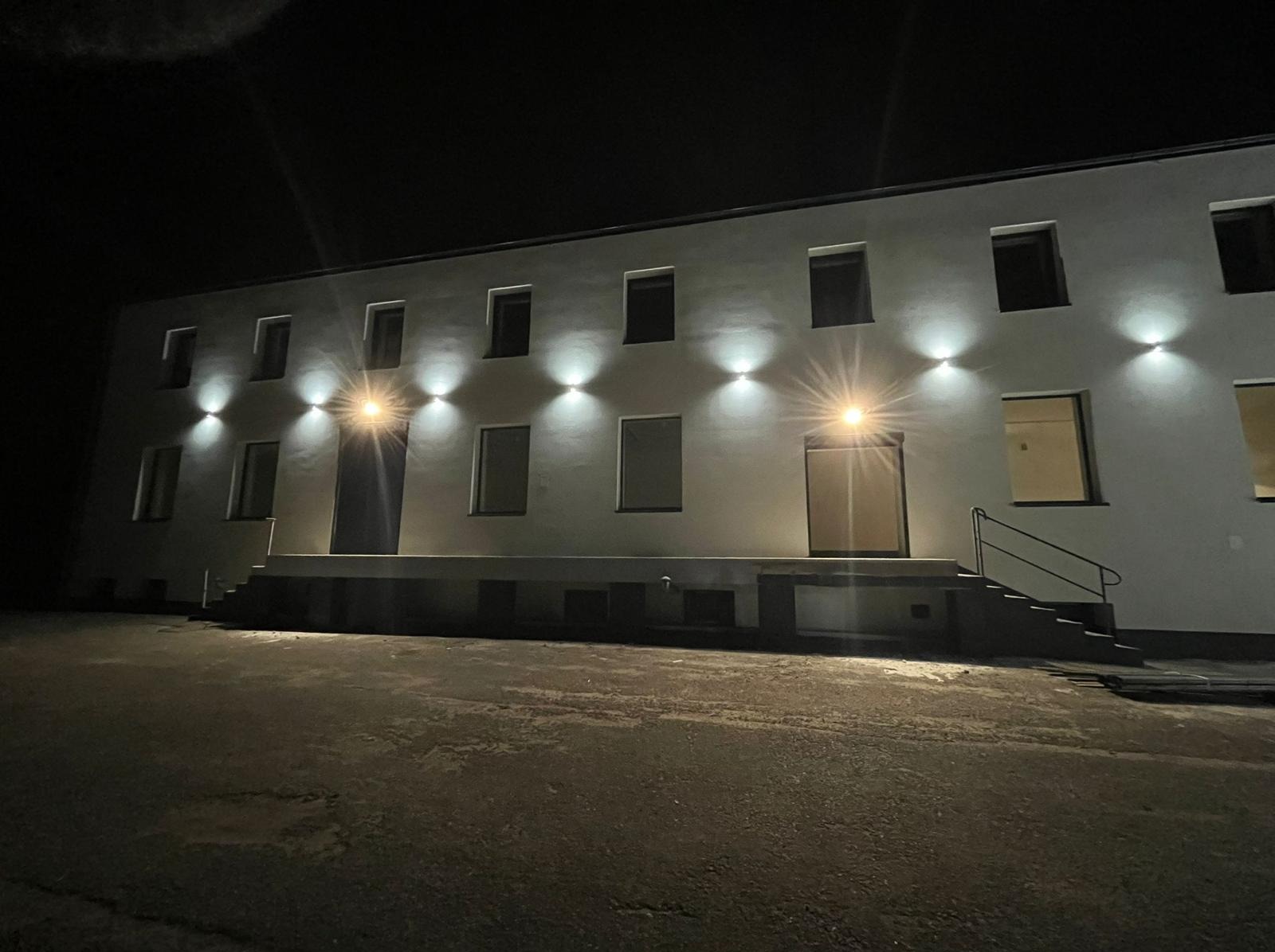 Oświetlony budynek nocą z równomiernie rozmieszczonymi lampami elewacyjnymi, tworzącymi efektowne stożki światła nad każdym oknem i drzwiami.