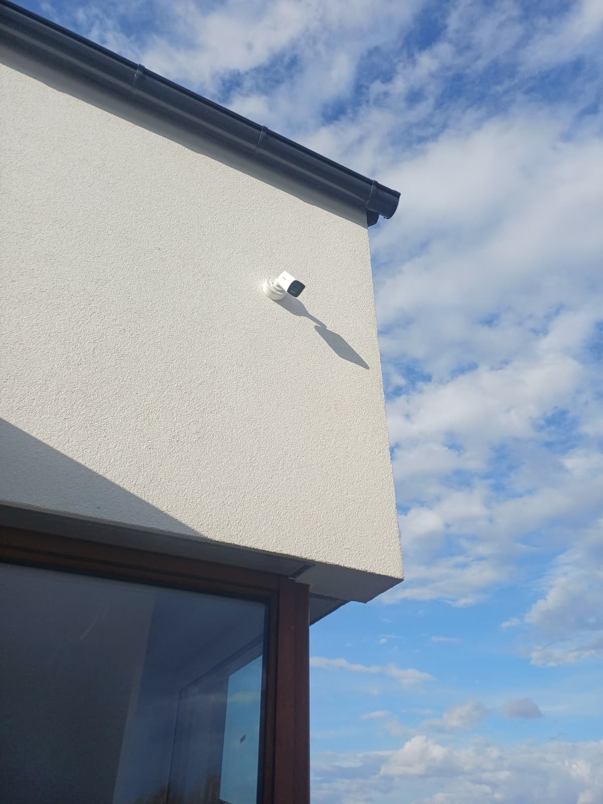 Biała kamera monitoringu zamontowana na elewacji budynku z widocznym oknem i fragmentem nieba z chmurami. Ujęcie z dołu, podkreślające nowoczesny design.