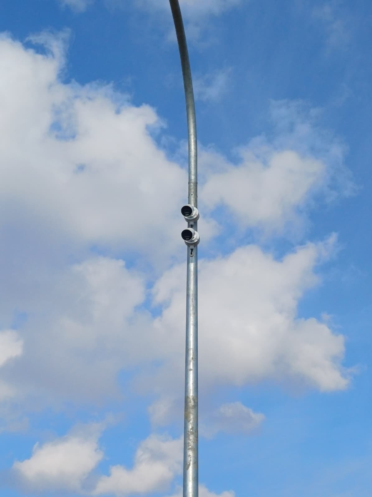 Metalowy słup oświetleniowy z dwoma reflektorami na tle błękitnego nieba z białymi obłokami. Widok z dołu, podkreślający wysokość konstrukcji.