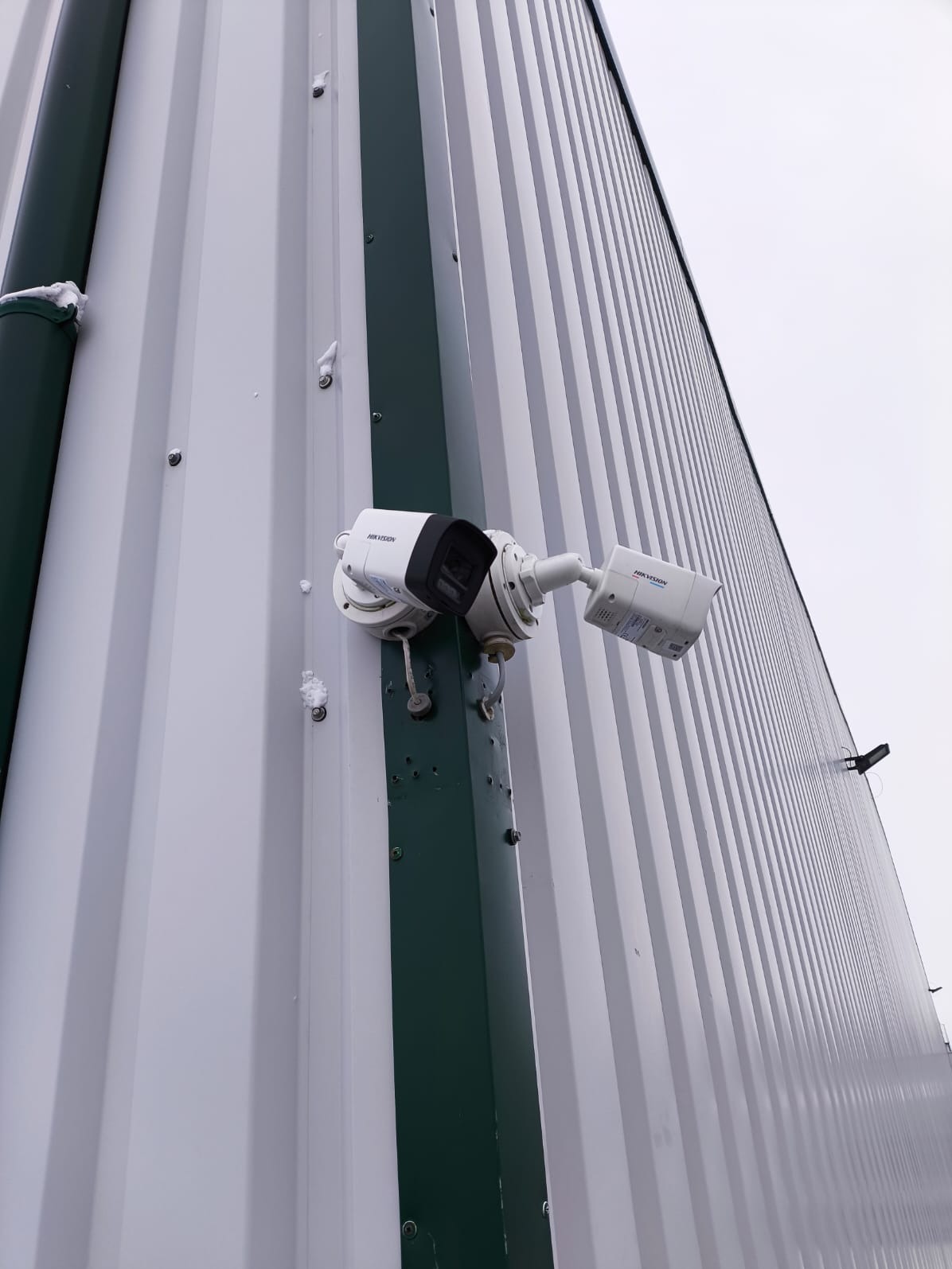 Dwie kamery monitoringu Hikvision zamontowane na metalowej ścianie budynku z widocznymi śladami montażu i fragmentami śniegu. Ujęcie z dołu, pochmurne niebo w tle.