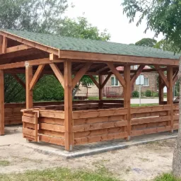 Nowa drewniana konstrukcja z zielonym dachem, przypominająca otwartą altanę z niskimi ściankami, ustawiona na betonowej podstawie w otoczeniu zieleni.