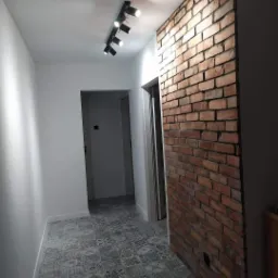 Korytarz po remoncie z dekoracyjną ścianą z czerwonej cegły, wzorzystą szaro-białą podłogą i oświetleniem punktowym na suficie.