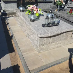 Granitowy nagrobek z nowo wyłożonym chodnikiem z kostki brukowej, otoczony piaskiem i ziemią.