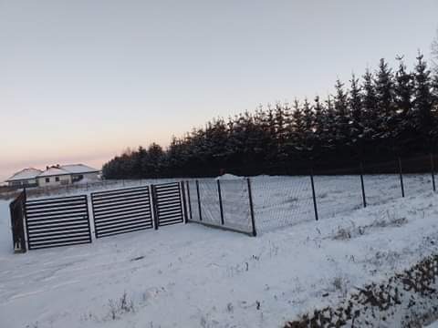 Zimowy krajobraz z nowym ogrodzeniem panelowym w kolorze antracytowym i siatką ogrodzeniową, na tle ośnieżonego pola i lasu iglastego, w oddali widoczne domy jednorodzinne.