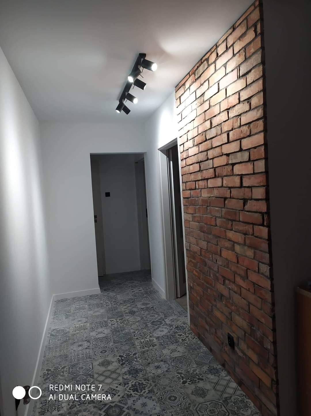 Korytarz po remoncie z dekoracyjną ścianą z czerwonej cegły, wzorzystą szaro-białą podłogą i oświetleniem punktowym na suficie.