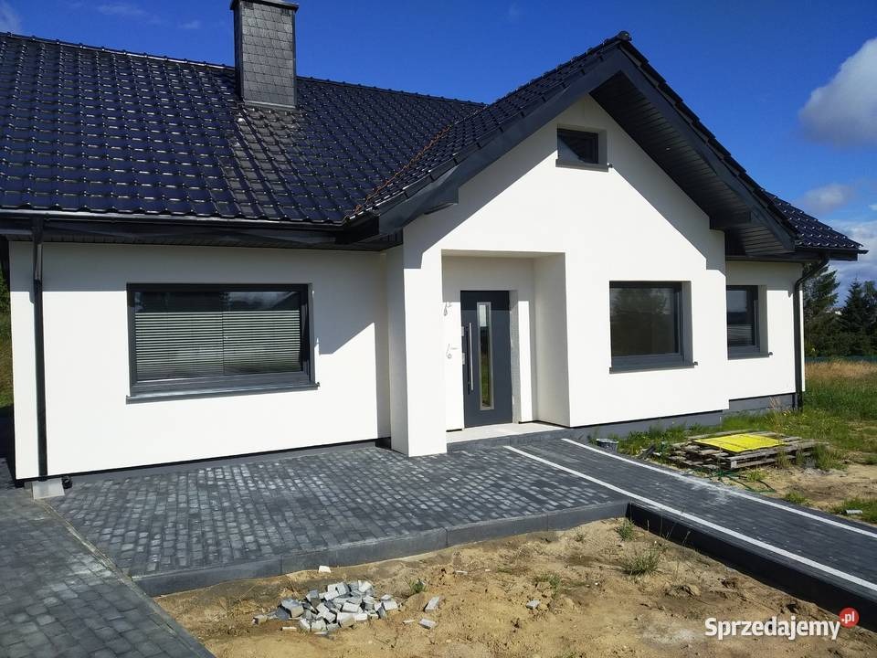 Nowoczesny dom jednorodzinny z białą elewacją, czarnym dachem z dachówki i szarą stolarką okienną, widok z zewnątrz na ułożoną kostkę brukową przed wejściem, z paletą i stertą kostki obok.