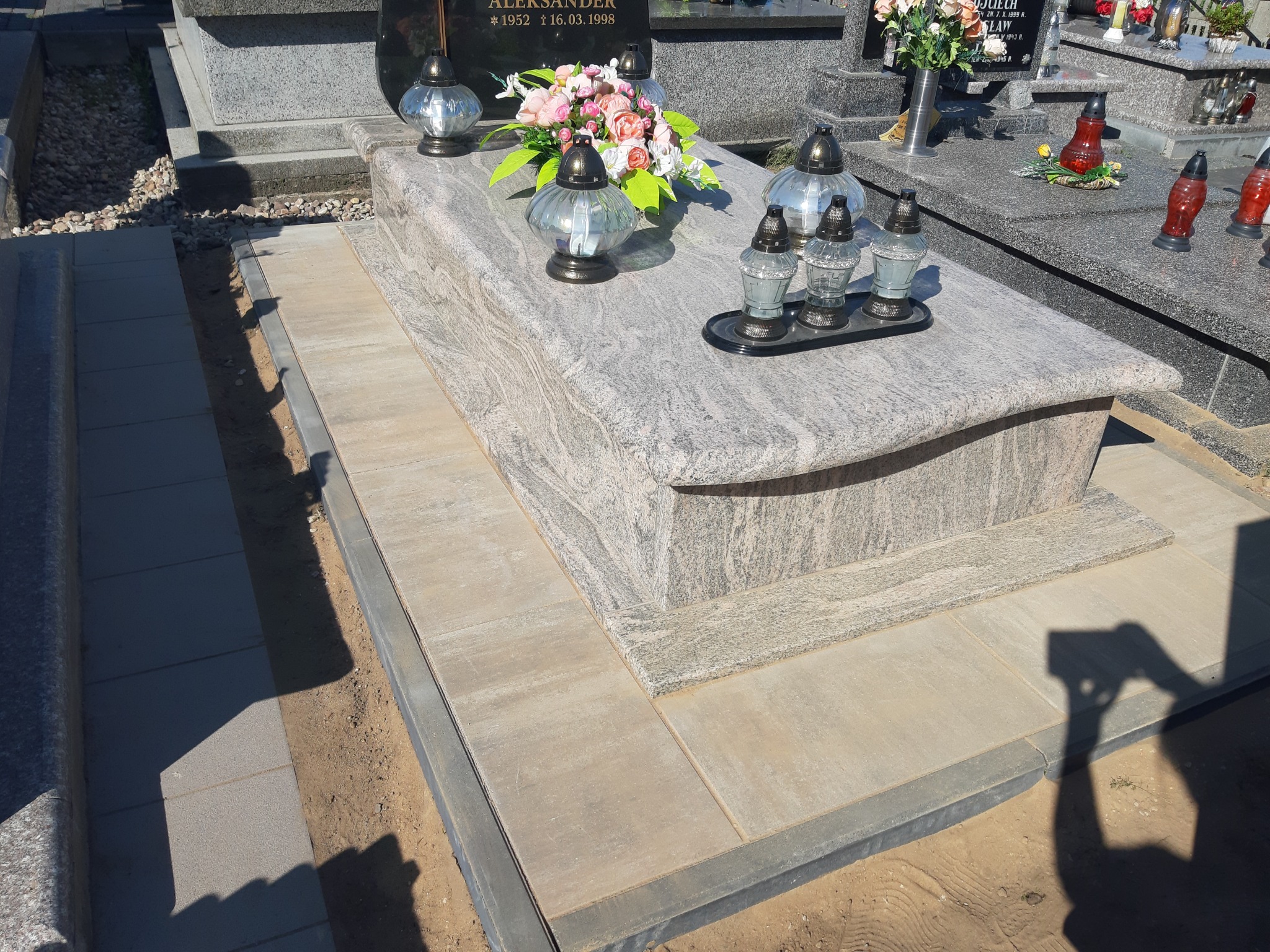 Granitowy nagrobek z nowo wyłożonym chodnikiem z kostki brukowej, otoczony piaskiem i ziemią.