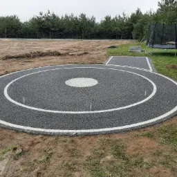 Okrągła powierzchnia z szarej kostki z białym obramowaniem w ogrodzie, widoczne metalowe pręty stabilizujące, w tle niebieski bus, trampolina i rząd drzew.