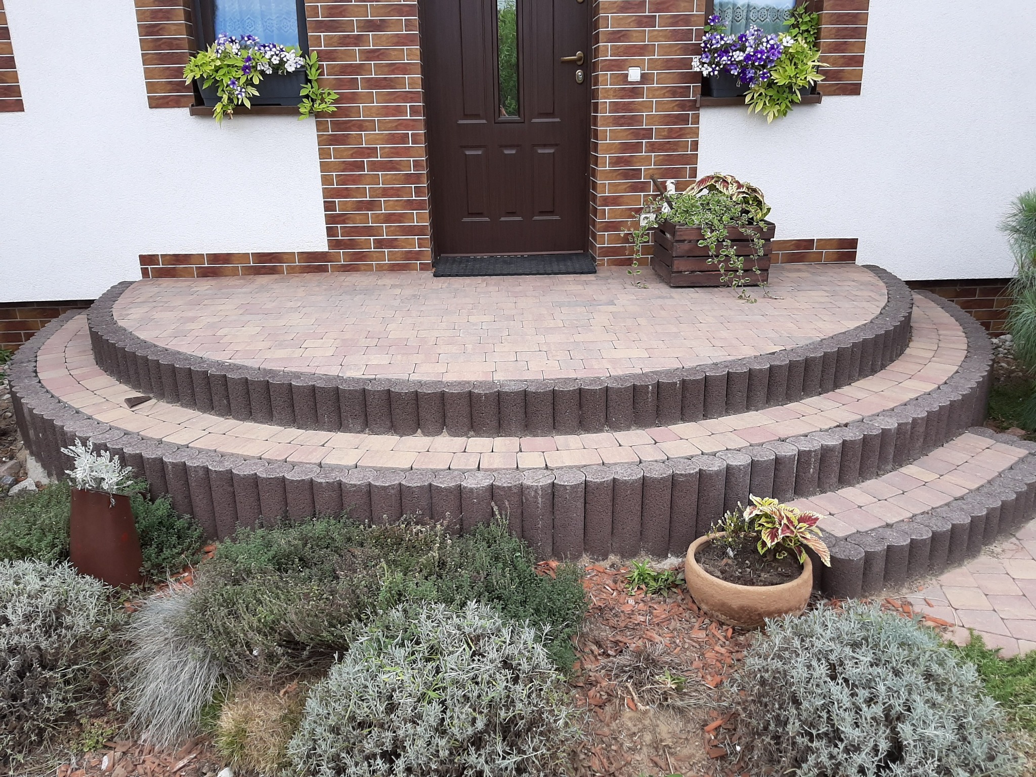 Trzy półokrągłe stopnie z kostki brukowej prowadzące do brązowych drzwi wejściowych, z roślinami w donicach i skrzynkach po bokach, widok z poziomu ogrodu.