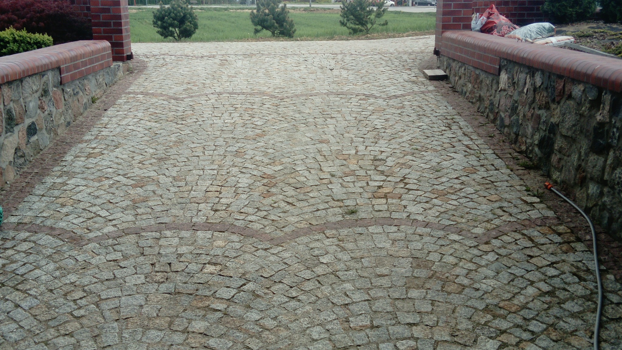 Ułożona z jasnoszarej kostki granitowej droga wjazdowa, z widocznymi fragmentami murków z kamienia i cegły po bokach oraz trawą w tle.