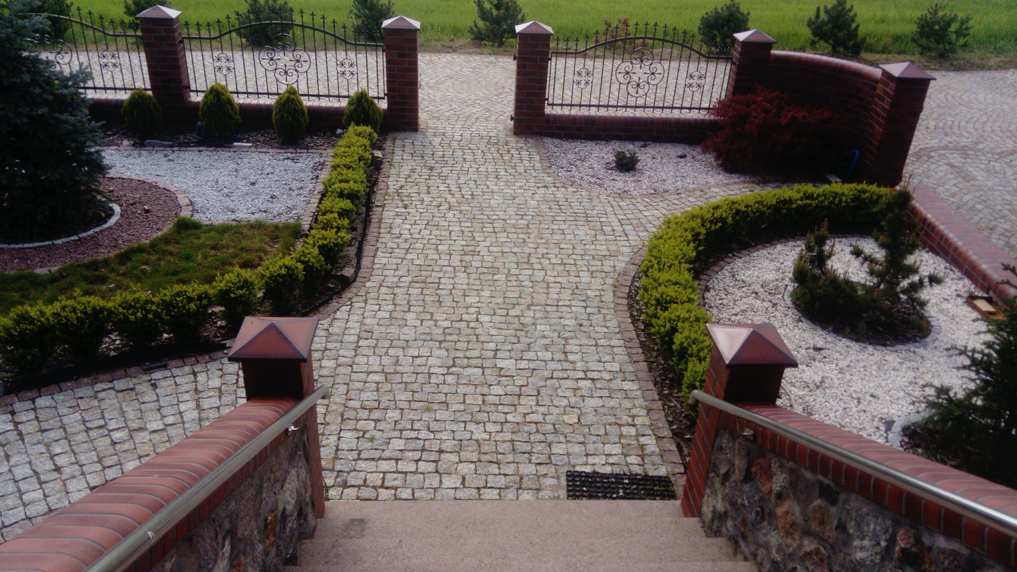 Widok z góry na ścieżkę z kostki granitowej prowadzącą przez ogród z niskimi krzewami, elementami z kamyków i ozdobnym ogrodzeniem z metalu i cegły.