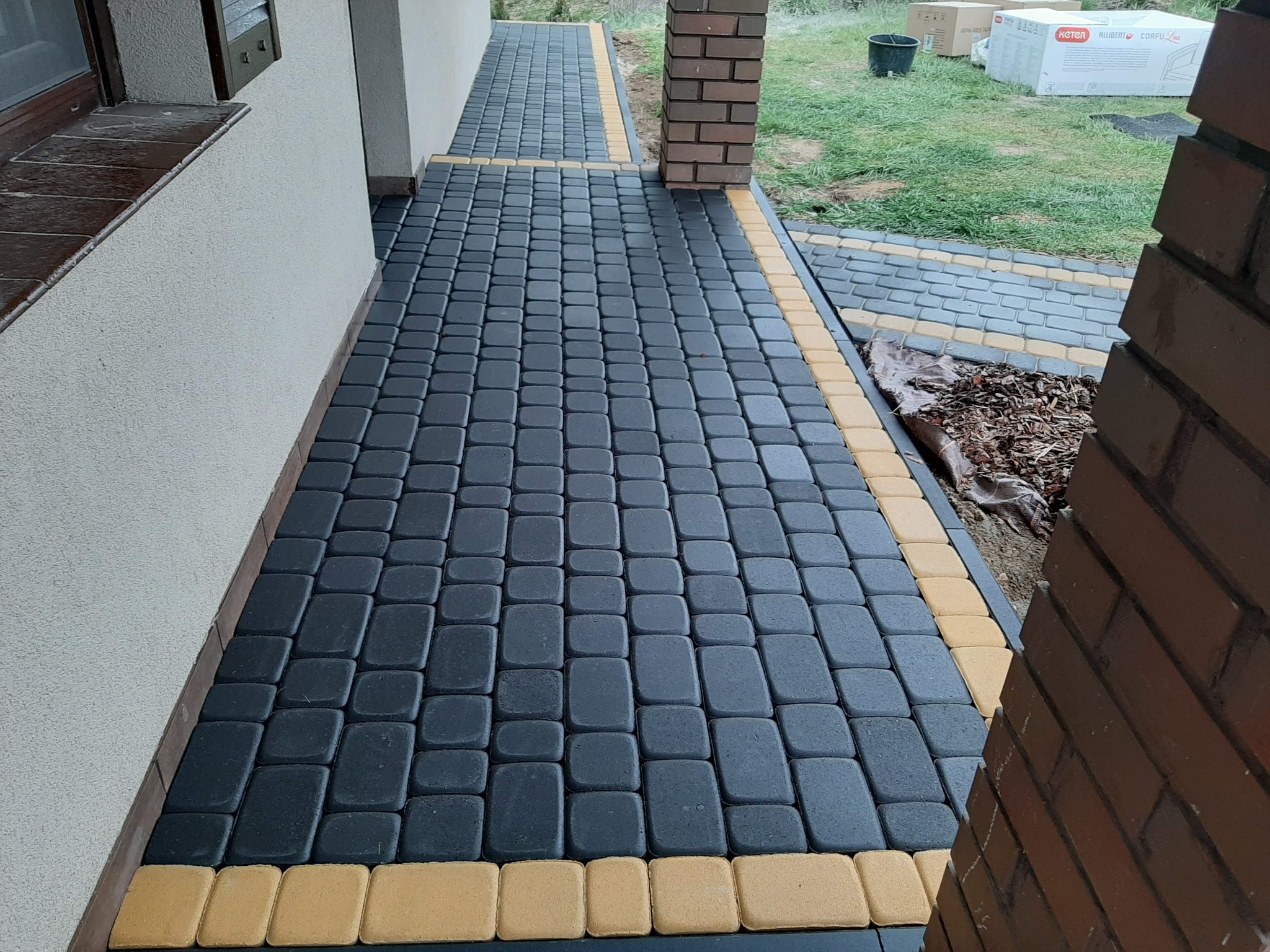 Ułożona nawierzchnia z ciemnoszarej kostki brukowej z obramowaniem z żółtej kostki, prowadząca wzdłuż ściany budynku i ceglanego filaru, z widocznymi kartonami w tle na trawniku.