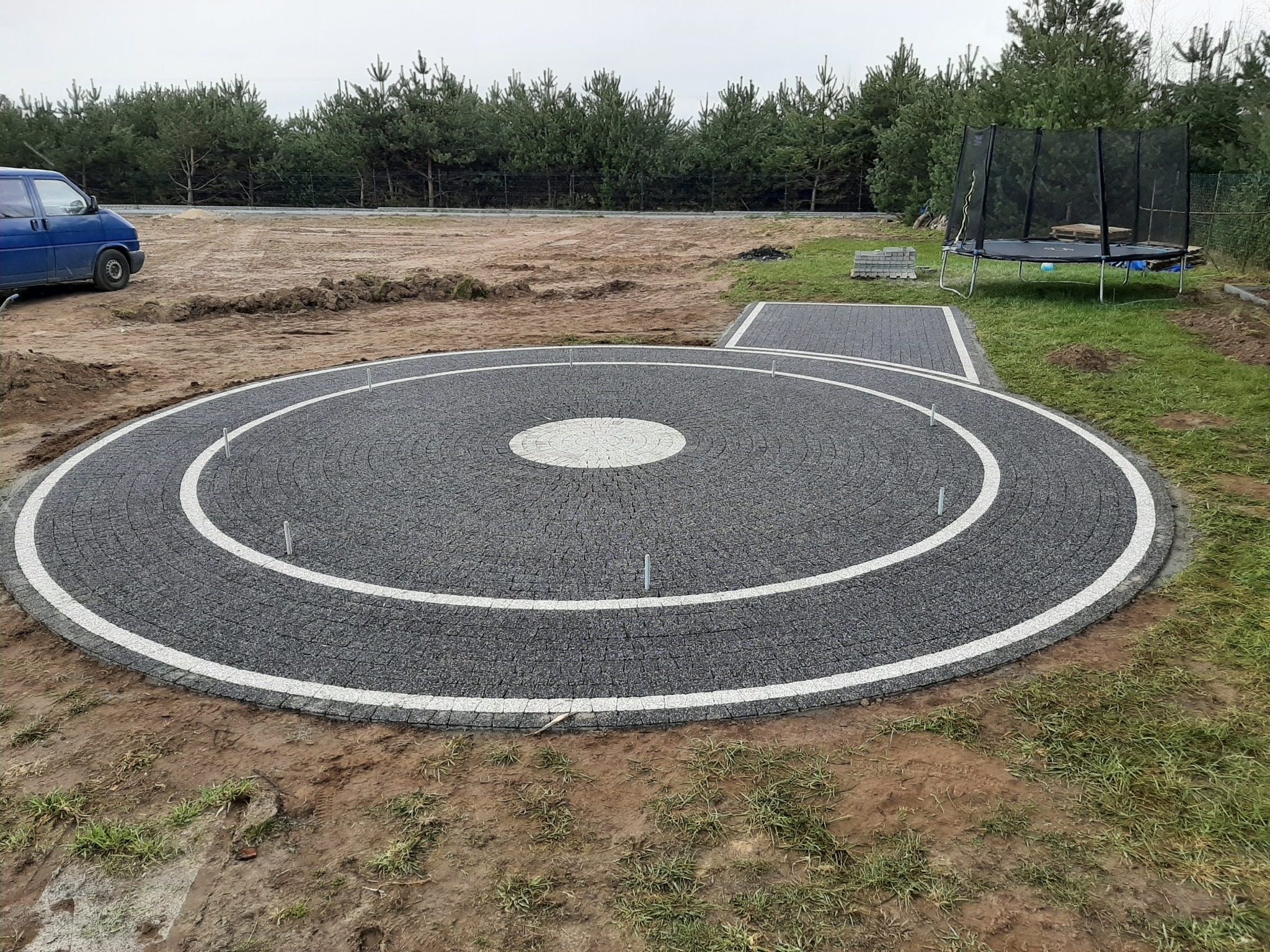 Okrągła powierzchnia z szarej kostki z białym obramowaniem w ogrodzie, widoczne metalowe pręty stabilizujące, w tle niebieski bus, trampolina i rząd drzew.