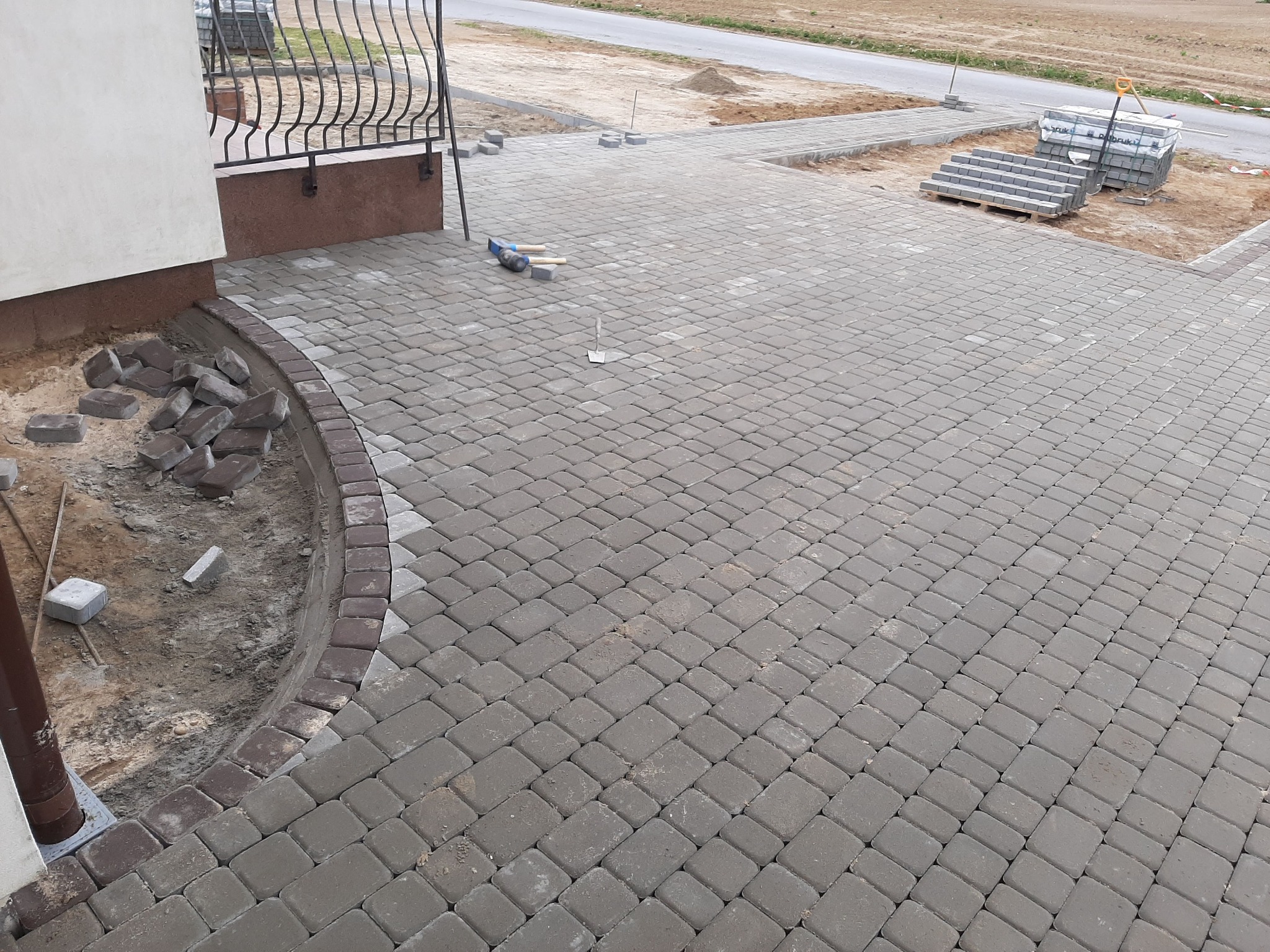 Fragment podjazdu z szarej kostki brukowej ułożonej w regularny wzór, z widocznymi narzędziami brukarskimi i paletami kostki w tle, obrzeża z ciemniejszej kostki przy budynku.