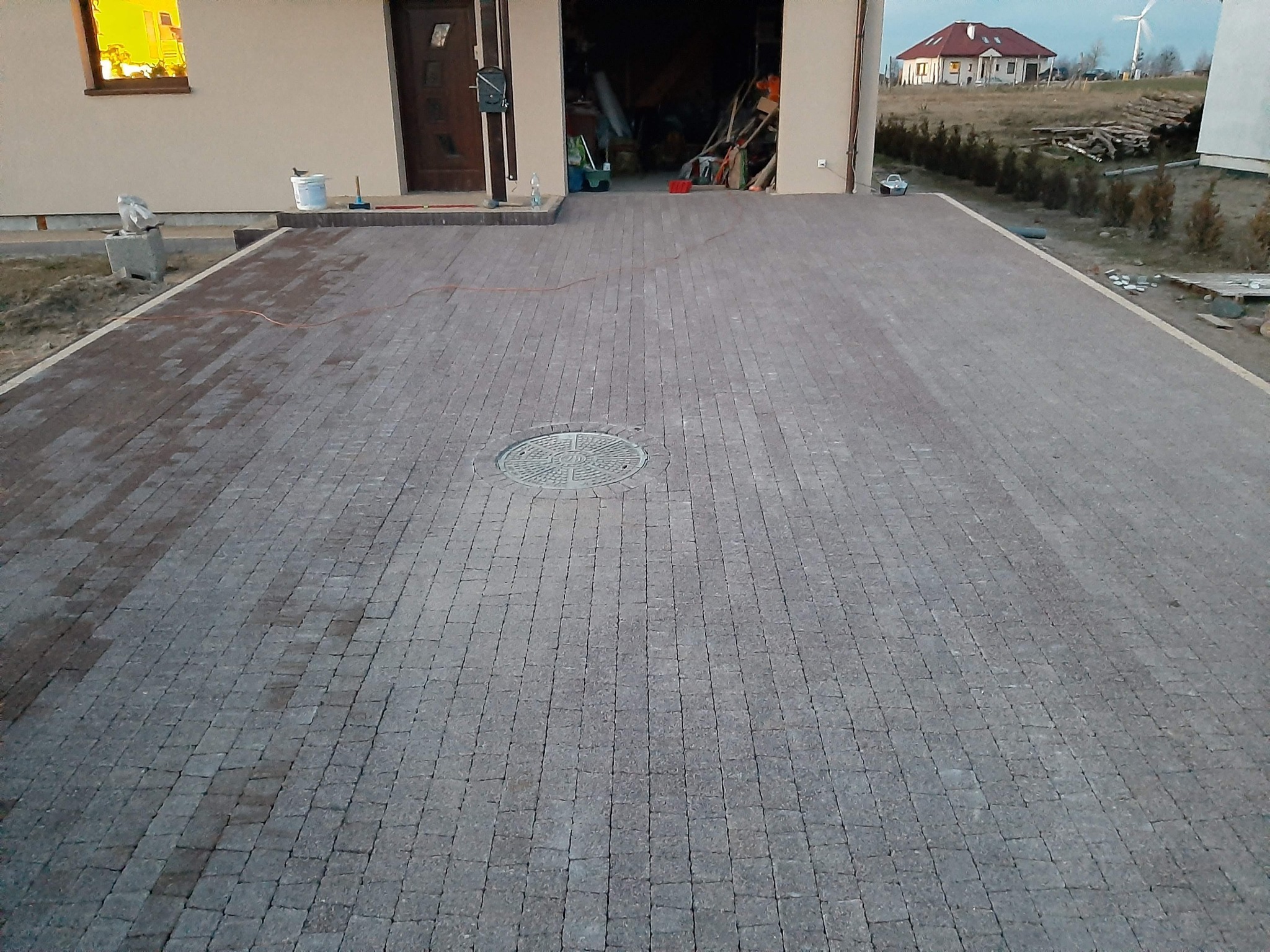 Ułożona nawierzchnia z kostki brukowej w odcieniach szarości i brązu, z okrągłą studzienką kanalizacyjną pośrodku, prowadząca do garażu domu jednorodzinnego z widocznym oknem i drzwiami wejściowymi...