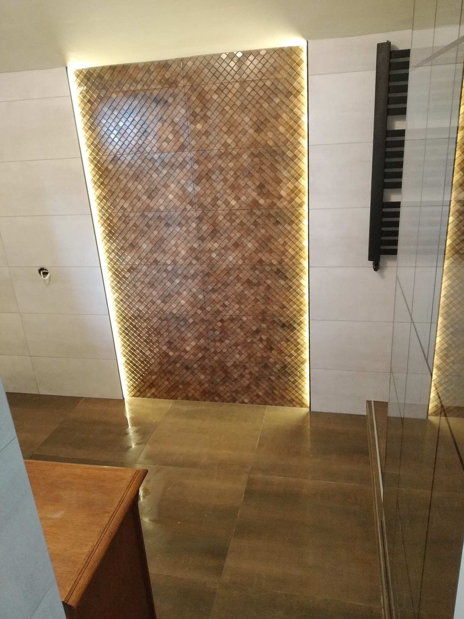 Wykończona ściana łazienki z dekoracyjną mozaiką drewnopodobną podświetloną listwą LED, obok grzejnik drabinkowy i szklana ścianka prysznicowa.