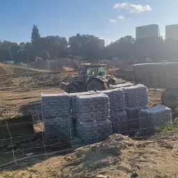 Stosy worków z cementem Cemex 32.5 R na terenie budowy, w tle traktor i wykopaliska ziemne, widoczne budynki mieszkalne w oddali.